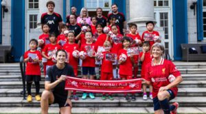 A group of smiling children in red Liverpool football shirts hold up footballs. His Majesty’s Ambassador to Japan, Julia Longbottom Kneels at the front with another women holding up a Liverpool Football Club scarf.