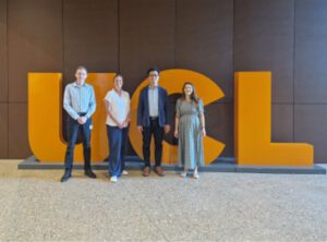 Four students stand in front of a large yellow UCL logo